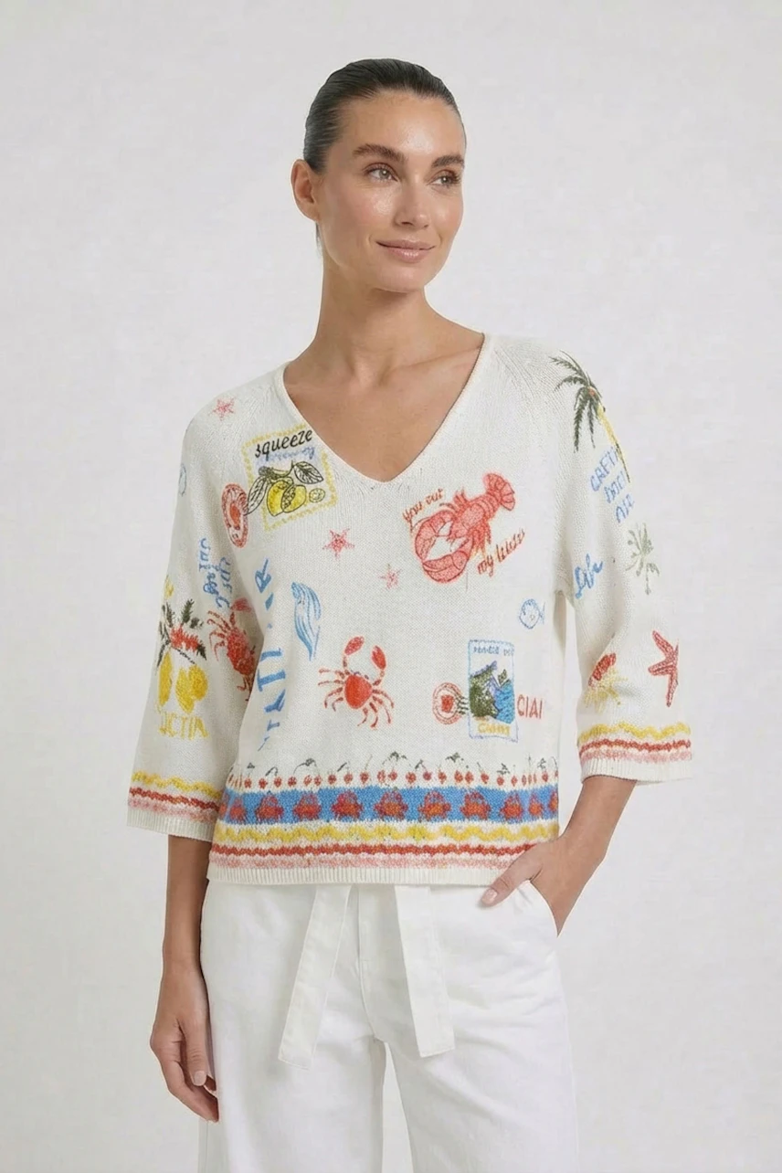 Frau im bunten Pullover mit maritimen Motiven und V-Ausschnitt, kombiniert mit weißen Hosen.