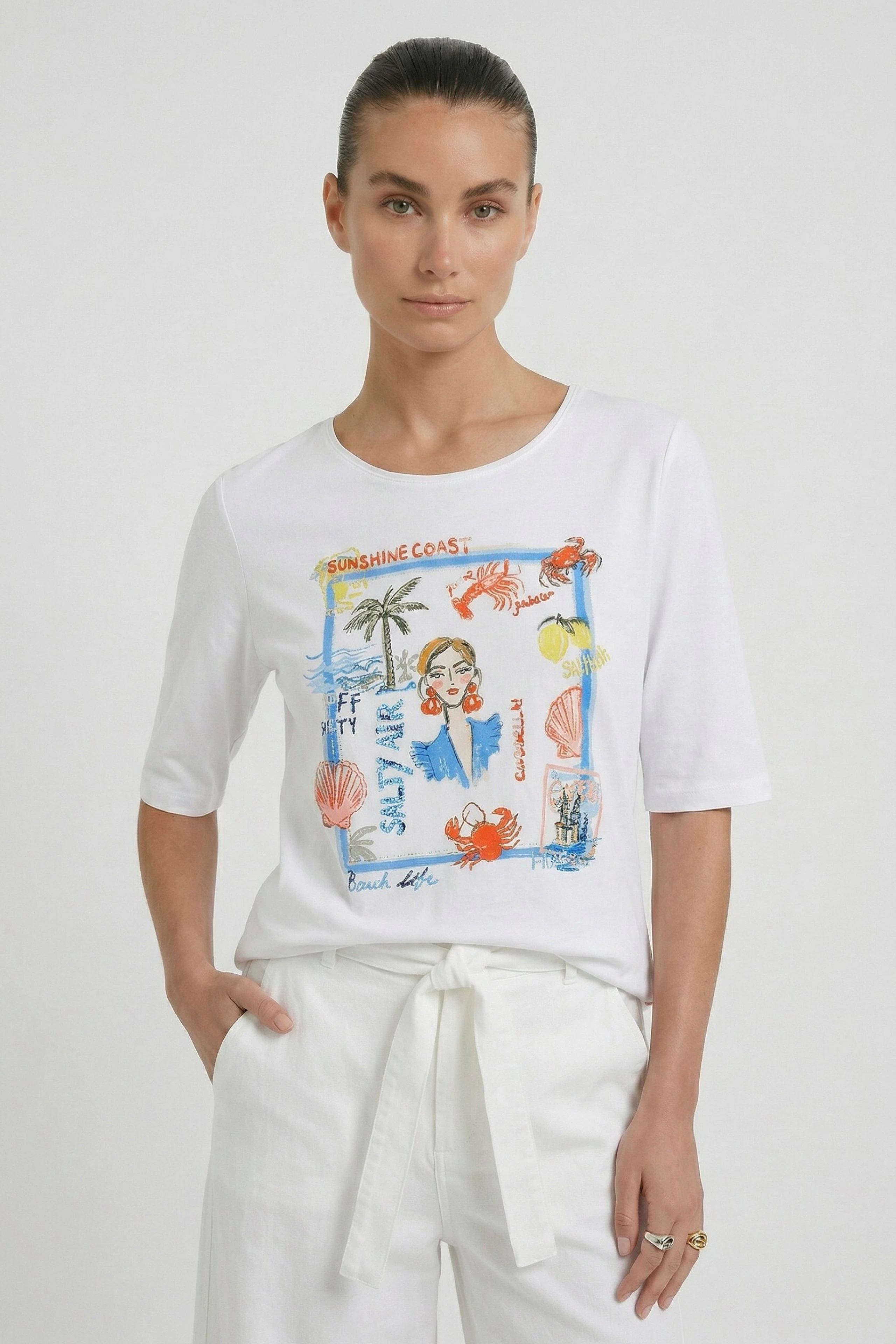 Casual women's T-shirt: Woman in a white T-shirt with colorful beach print, paired with white pants.