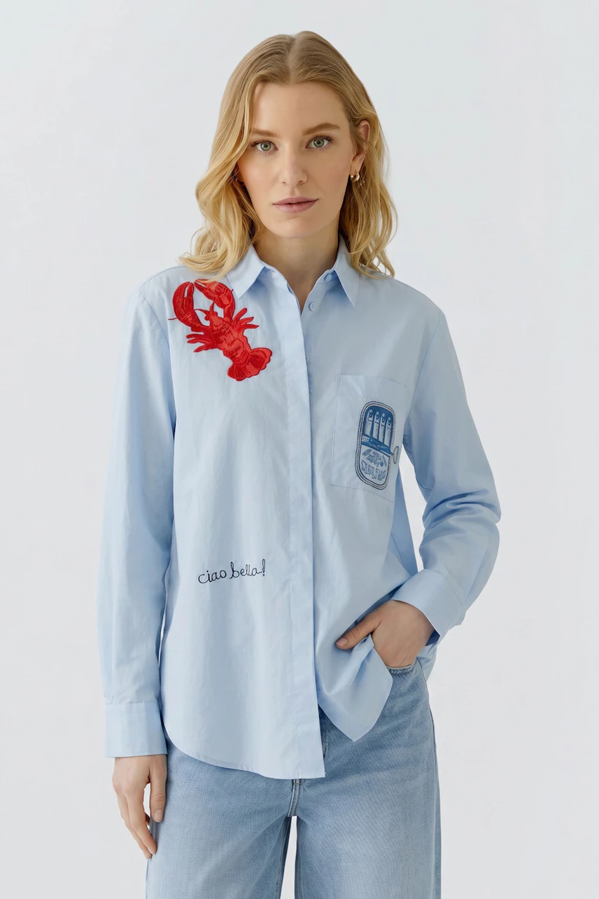 Lässige Damen-Bluse: Frau in hellblauer Bluse mit rotem Hummer-Print und Schriftzug, kombiniert mit weißen Hosen.