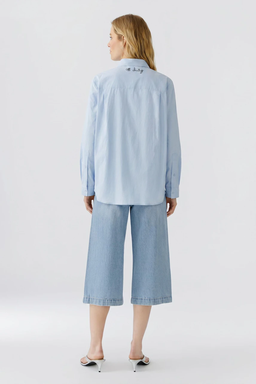 Lässige Damen-Bluse: Frau in hellblauer Bluse und weißen Hosen, von hinten gesehen.