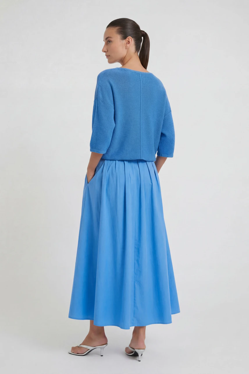 Woman in a blue midi skirt and matching top, standing sideways, hands in pockets, wearing white heels.