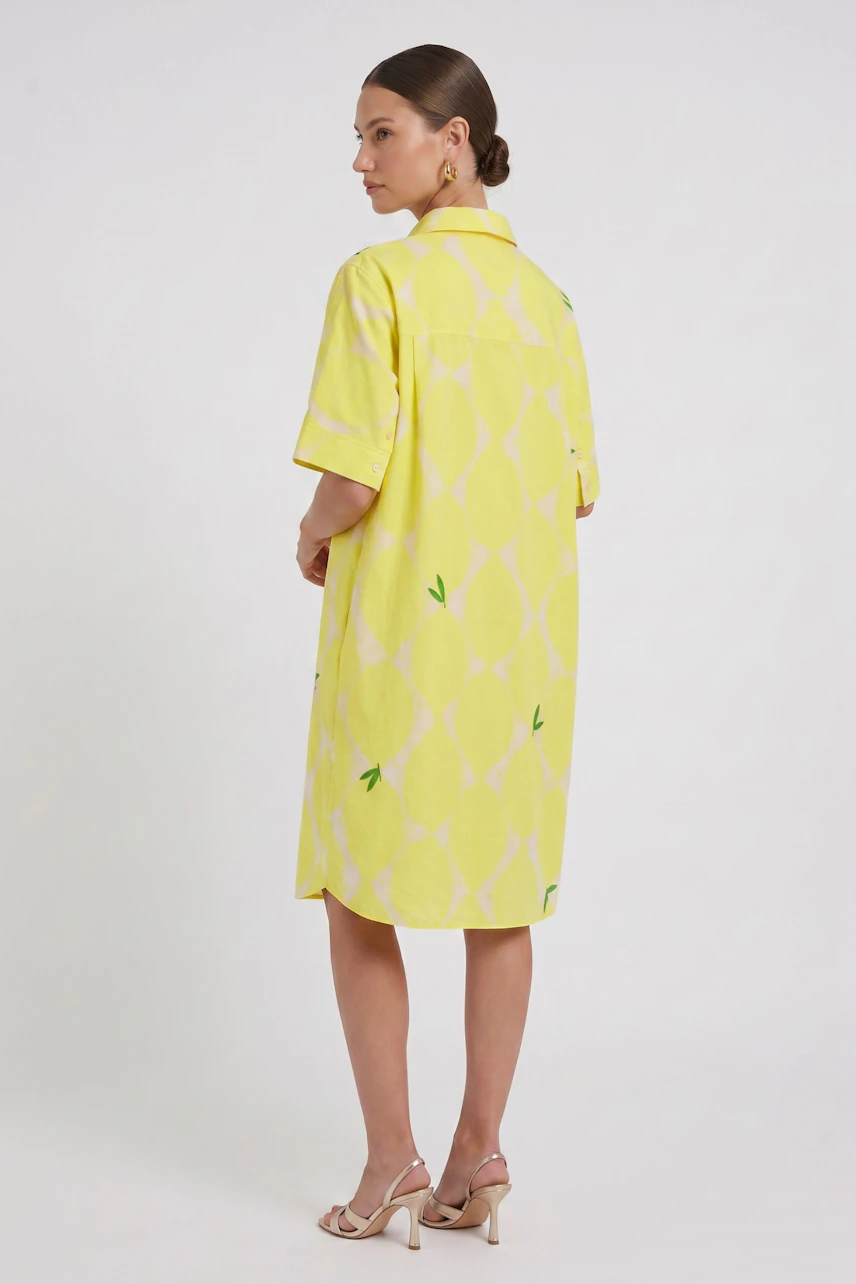 Frau im gelben Hemdblusenkleid mit Muster, seitlich stehend, zeigt Rückansicht. Sommerlich und lässig.