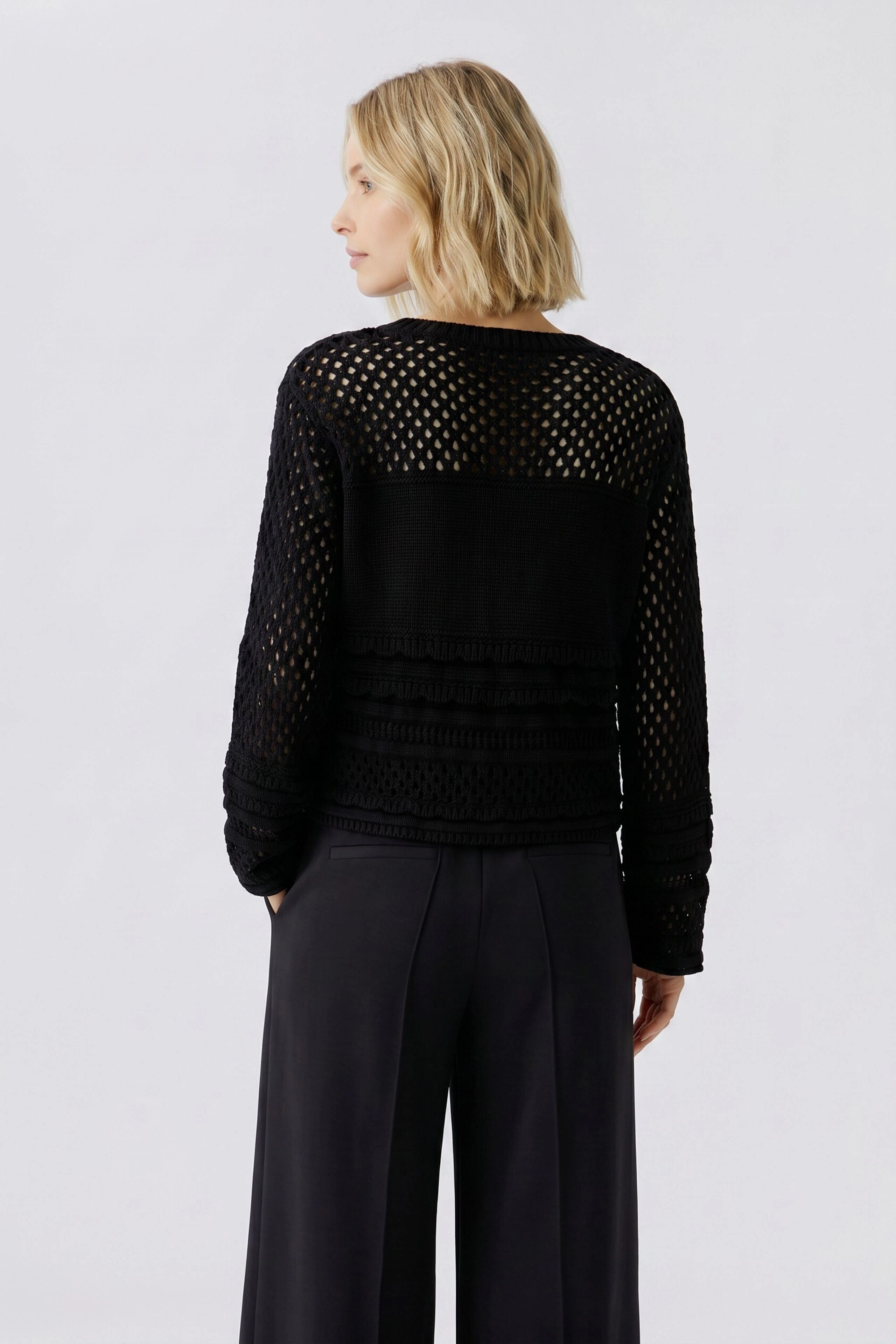Frau im schwarzen Pullover mit Netzstruktur und Pailletten, kombiniert mit weiter Hose, von hinten gezeigt.