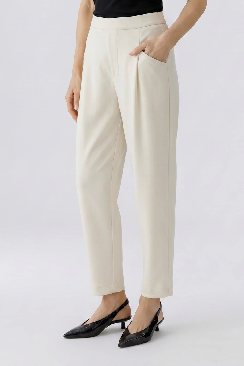 Woman in cream trousers, black top, and heels. Hands in pockets, showcasing casual style.