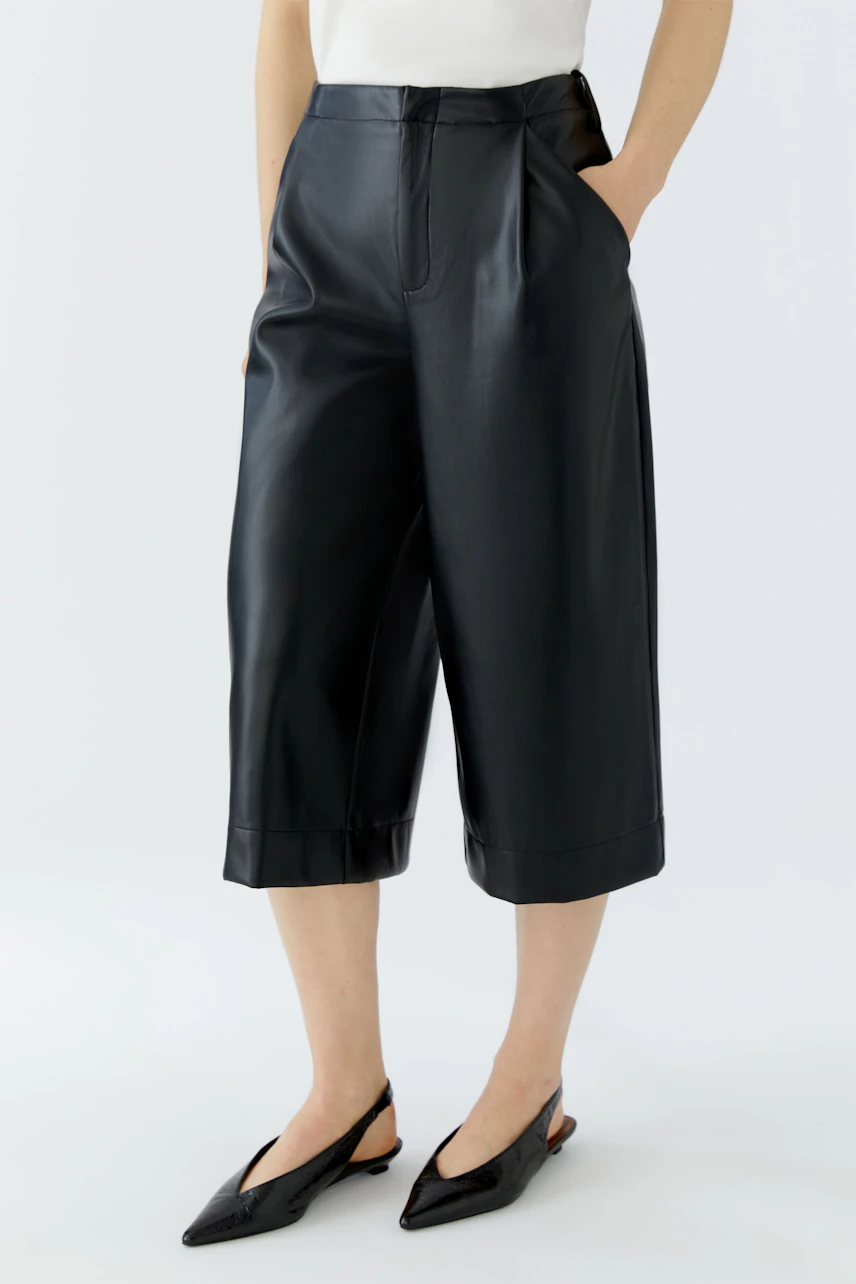 Woman in black pleated trousers, white top. Cropped length, hands in pockets, casual style.
