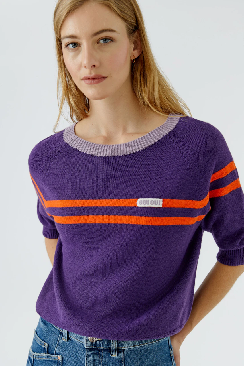 Lässiger Damen-Pullover: Frau im lila Pullover mit orangefarbenen Streifen und Jeans schaut entspannt.