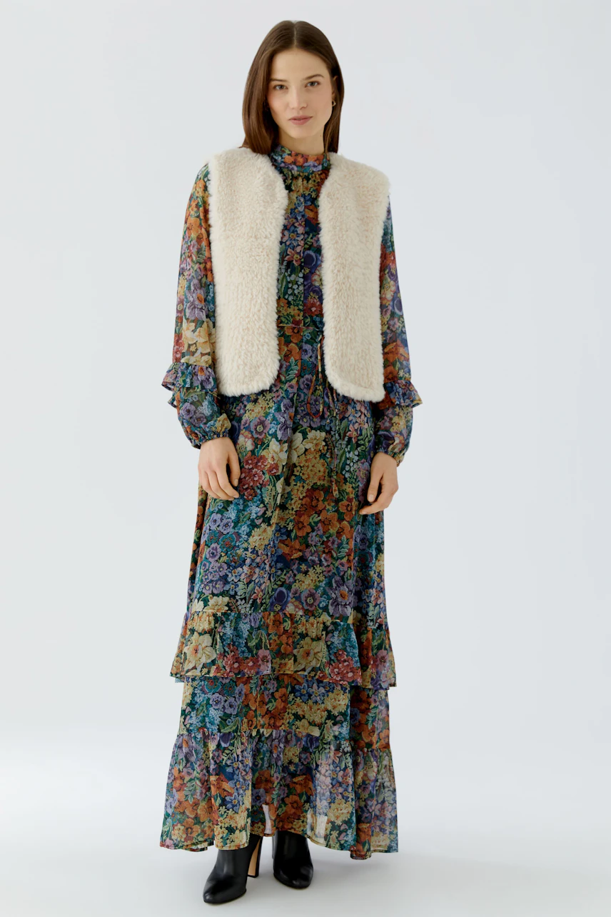Woman in floral dress with a white knit vest, standing confidently. Casual and stylish look.