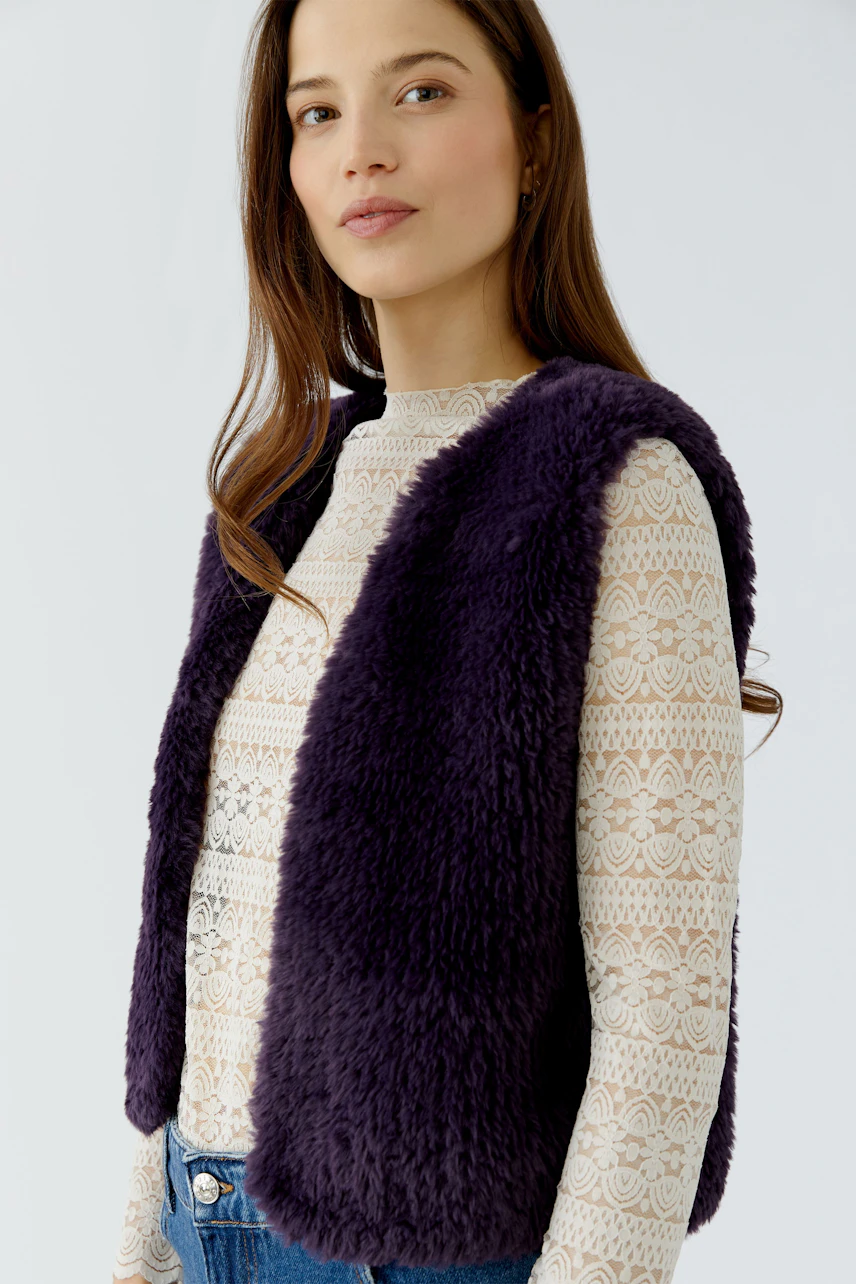 Woman in a purple knit vest over a lace top, paired with blue jeans, looking confidently.