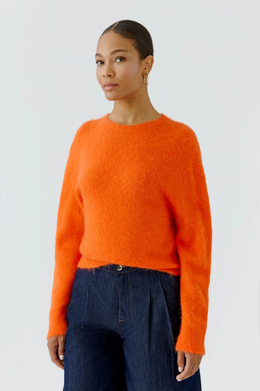 Frau im leuchtend orangefarbenen Pullover, kombiniert mit dunkelblauer Hose, schaut selbstbewusst.