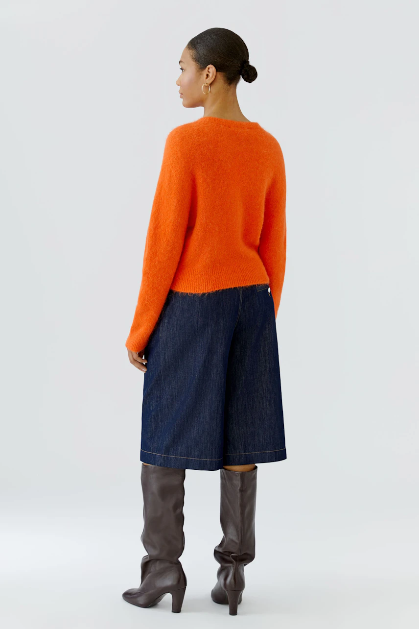 Frau im orangefarbenen Pullover, kombiniert mit Jeans-Culottes und braunen Stiefeln, von hinten gesehen.