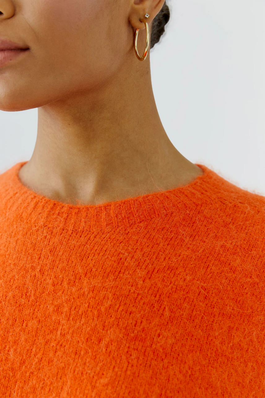 Frau im leuchtend orangefarbenen Pullover, schlichter Schnitt, goldene Ohrringe, dezentes Styling.