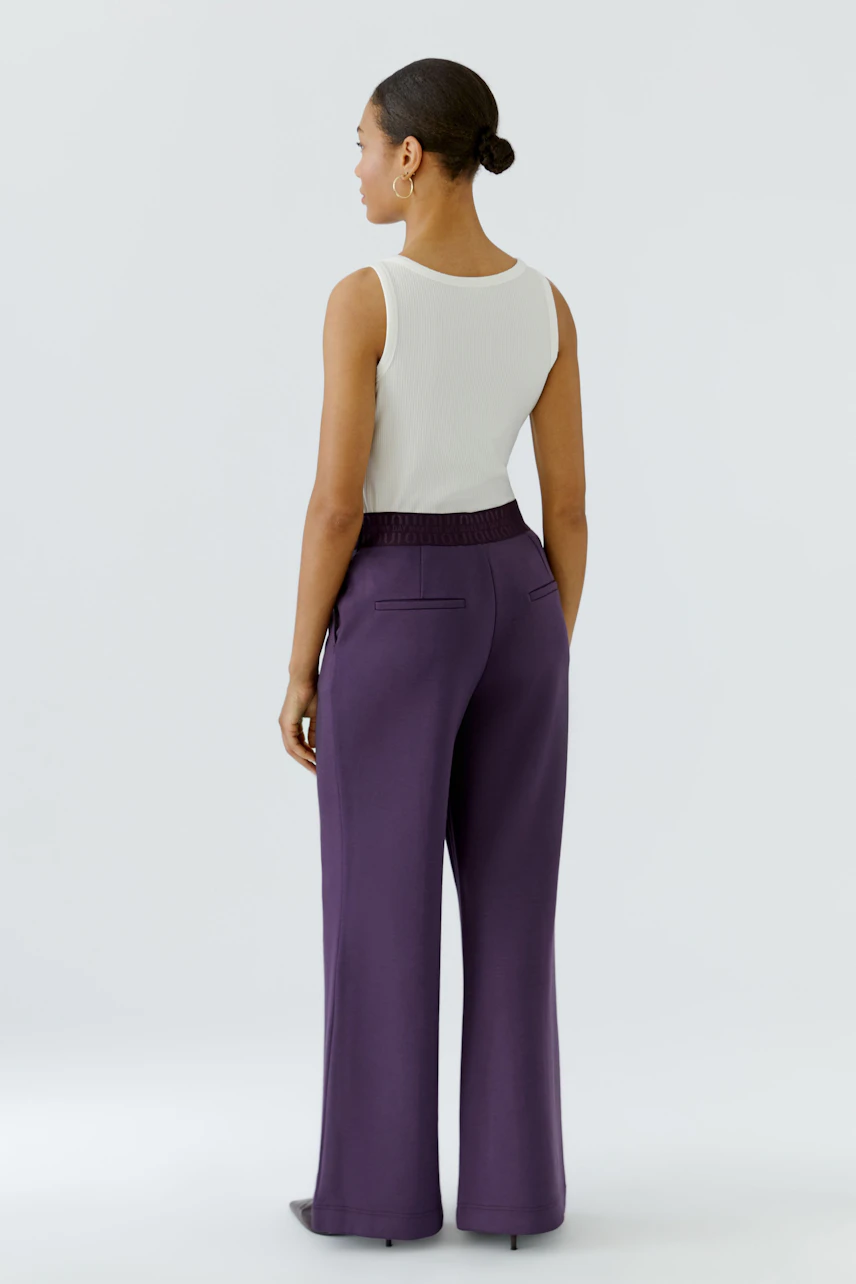 Woman in purple pants and white top, standing confidently. The pants have a relaxed fit and elastic waistband.
