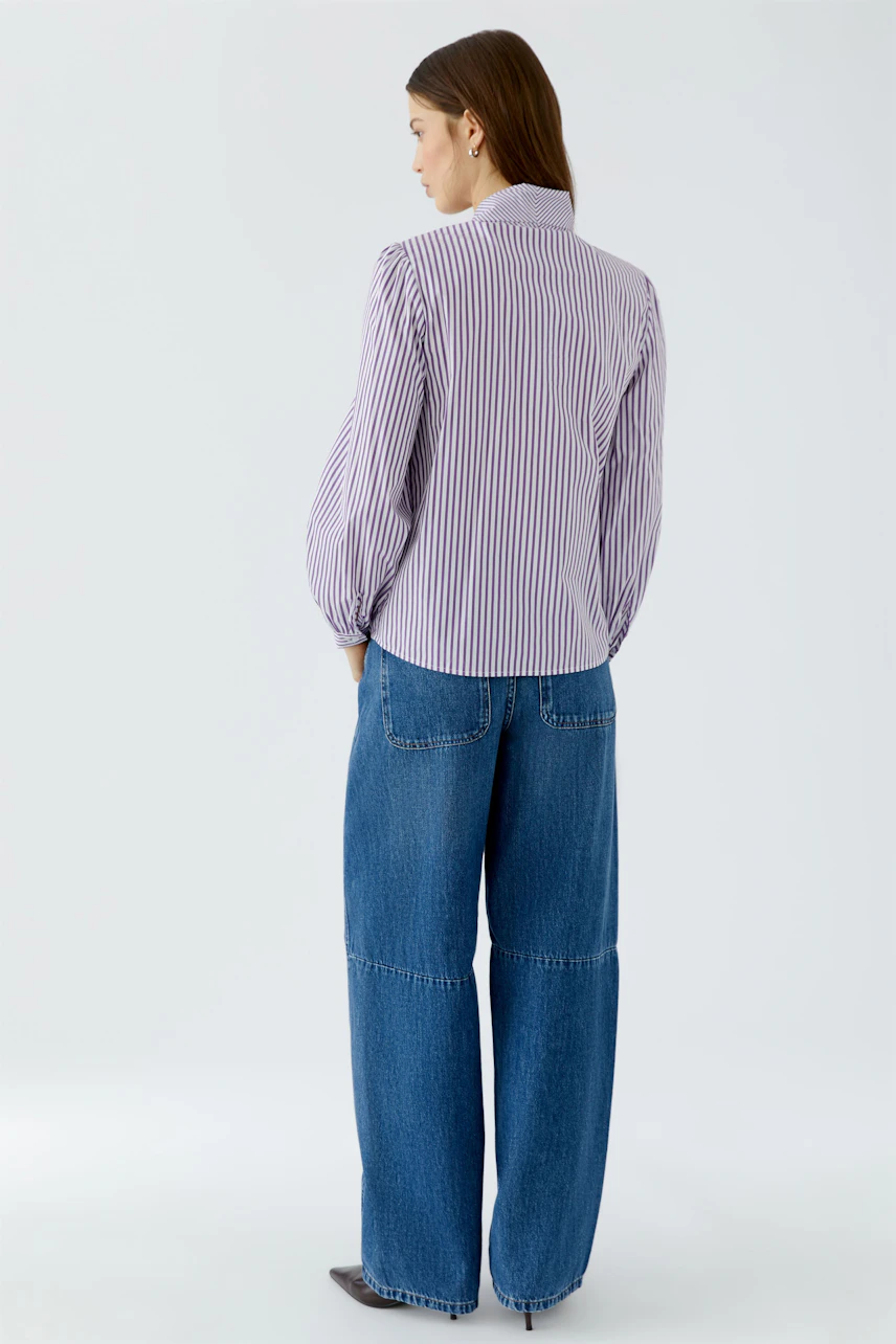 Frau in gestreifter Schluppenbluse und blauer Jeans, seitlich stehend, zeigt lässigen Look.