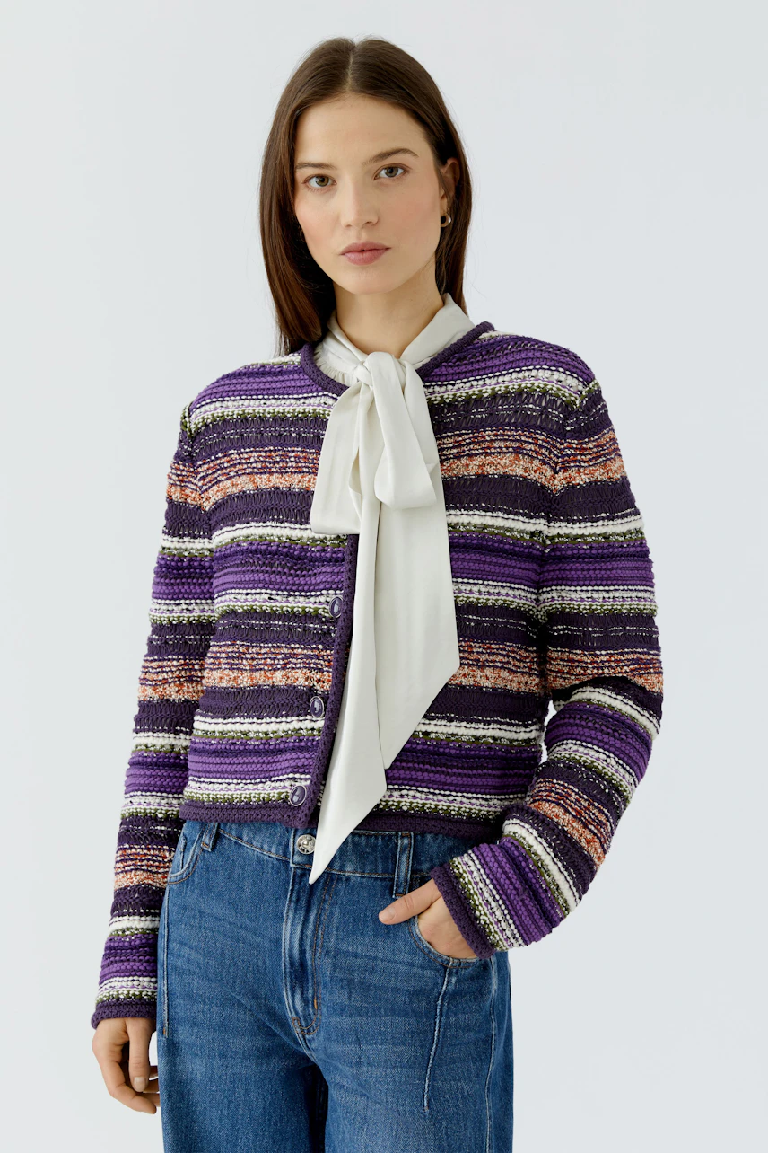 Frau in bunter Strickjacke mit Streifenmuster, kombiniert mit weißer Bluse und blauer Jeans.