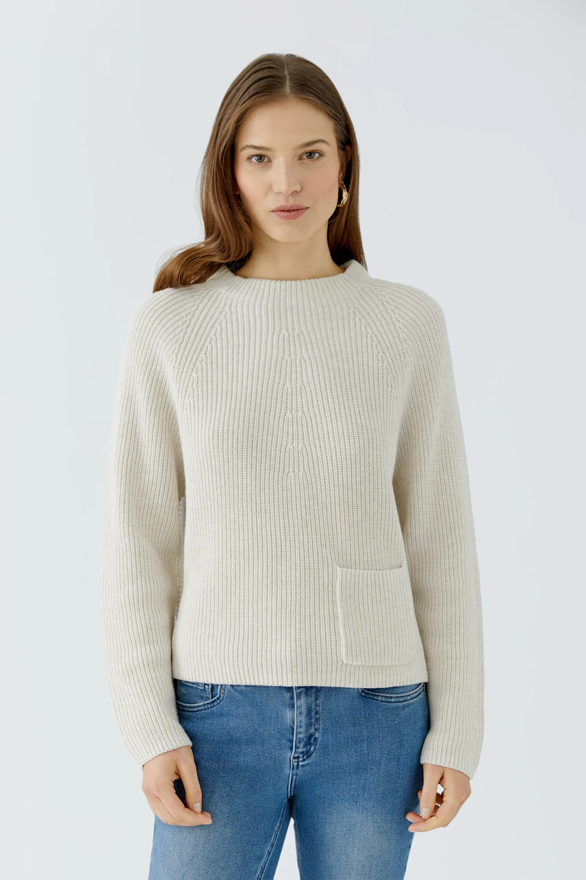 Frau im cremefarbenen Pullover, schlichtes Design, kombiniert mit blauer Jeans, schaut direkt in die Kamera.