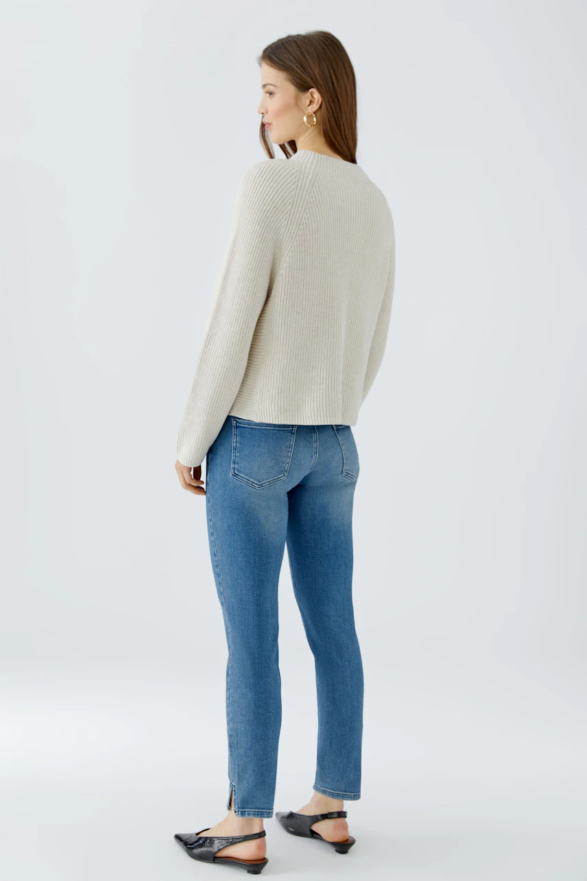 Damen-Pullover: Frau im cremefarbenen Pullover und blauer Jeans, seitlich stehend, mit schlichten schwarzen Schuhen.