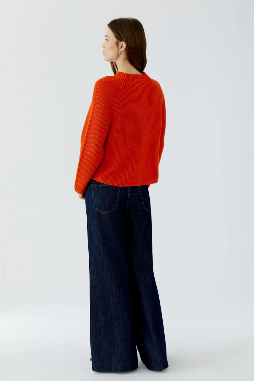 Frau im roten Pullover und blauer Jeans, seitlich stehend. Lässiger Look, ideal für den Alltag.