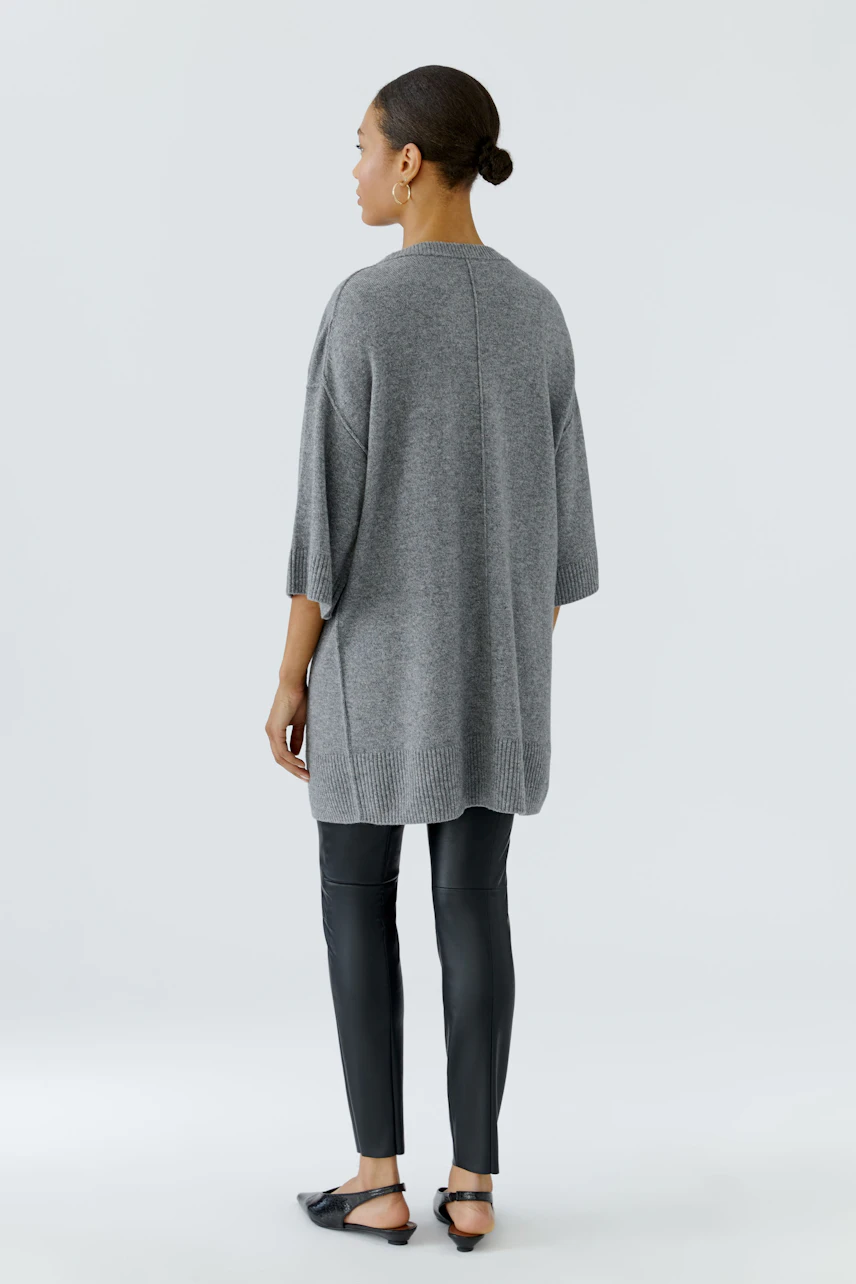 Casual women's pullover: Woman in a loose grey pullover with black leggings and sandals, viewed from the back.