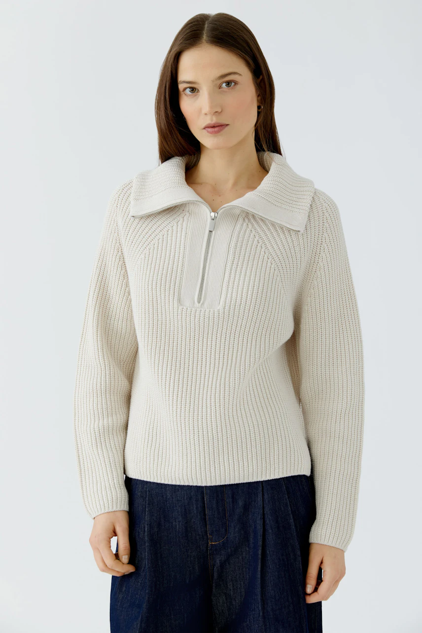 Frau im cremefarbenen Pullover mit Reißverschluss, kombiniert mit dunkler Jeans. Schlicht und modern.