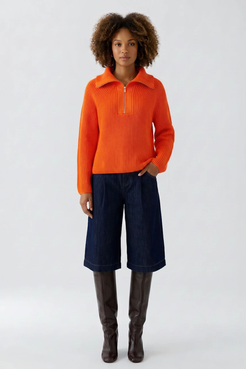 Woman in vibrant orange pullover, paired with denim culottes and knee-high boots, standing confidently.