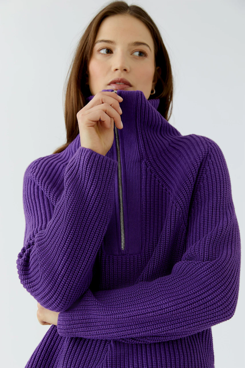 Casual women's pullover: Woman in a purple knit pullover with a high collar, looking thoughtful.