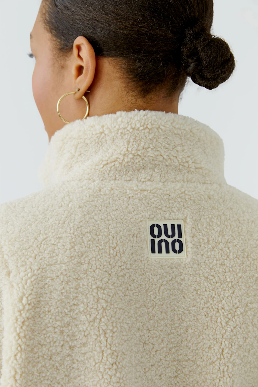 Woman in cozy blouson with soft texture, logo on back, hair in bun, wearing hoop earrings.