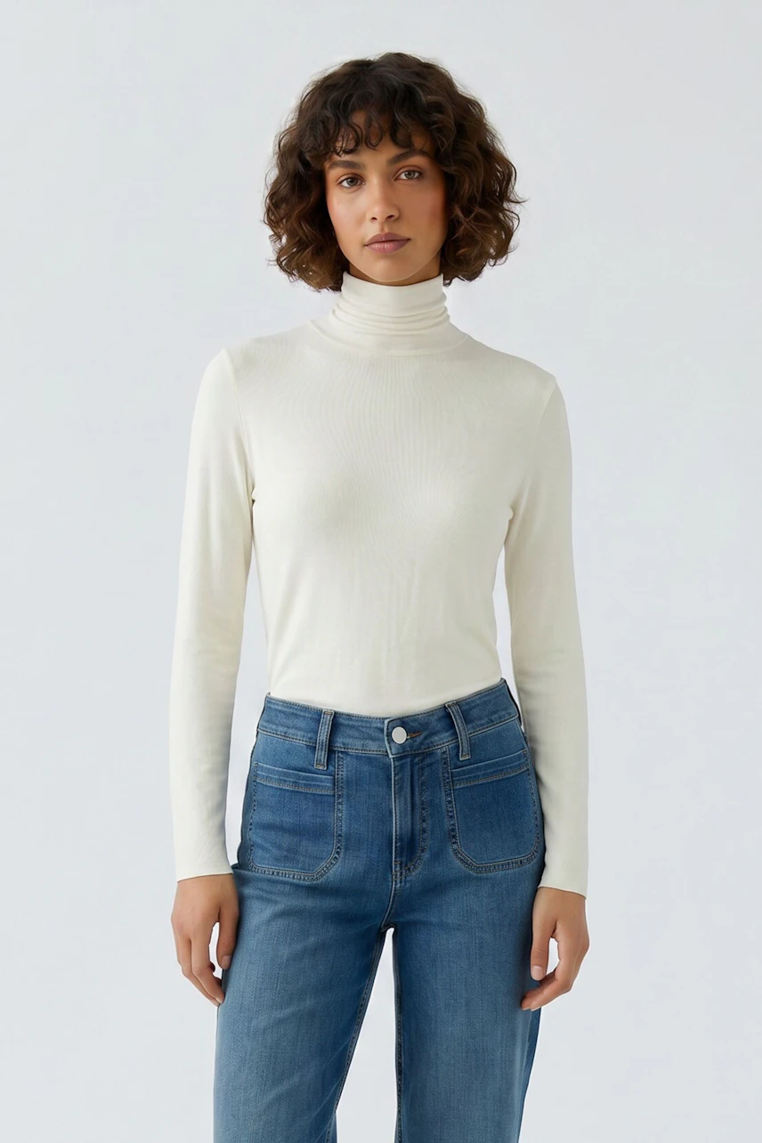 Frau im weißen Langarmshirt mit Rollkragen, kombiniert mit blauer Jeans, schaut selbstbewusst.