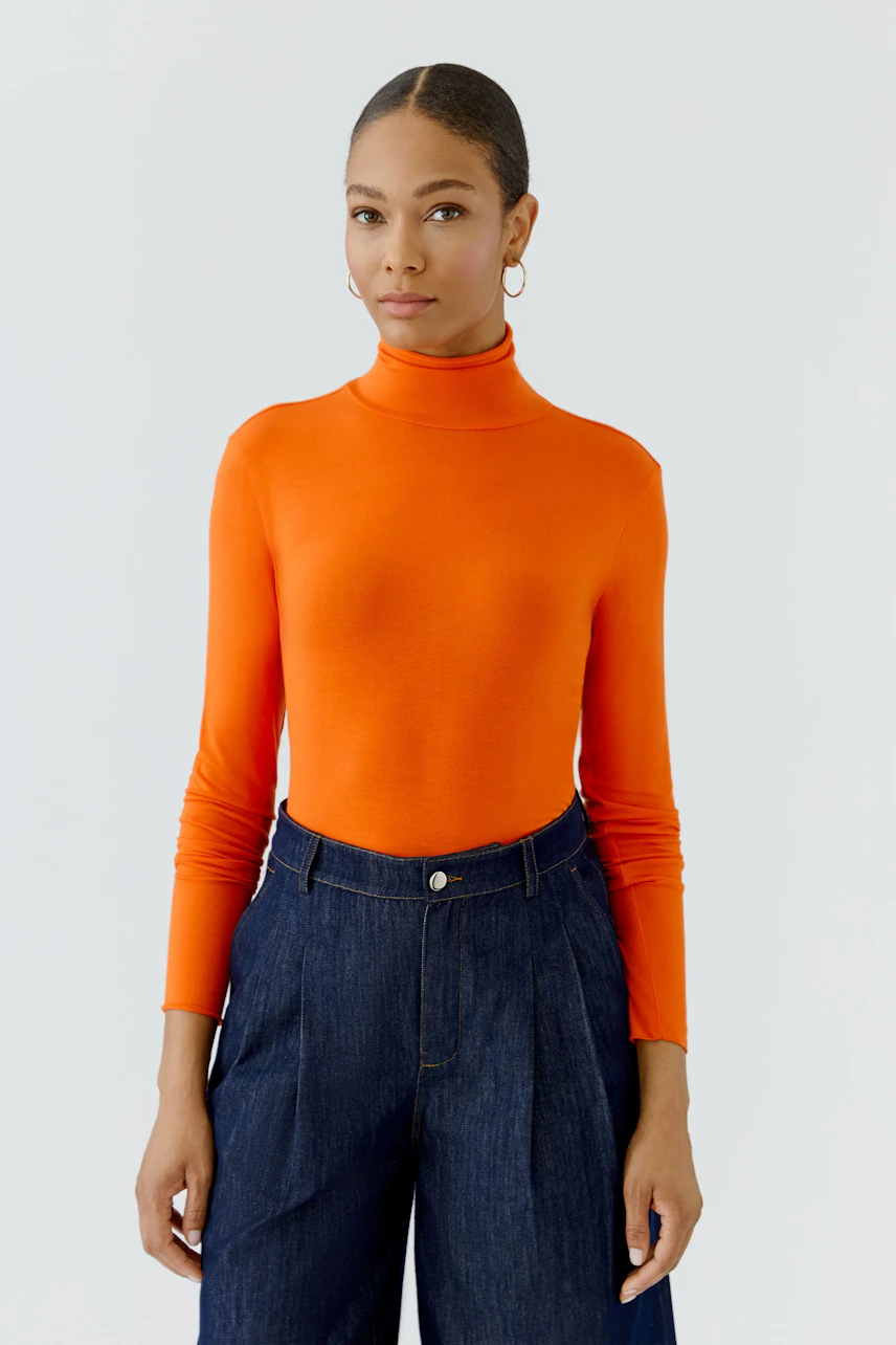 Woman in vibrant orange long-sleeve shirt, paired with high-waisted denim pants, standing confidently.