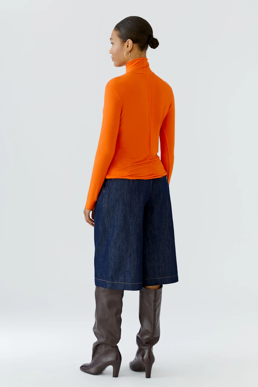 Woman in orange long-sleeve shirt, denim culottes, and brown boots, viewed from the back.