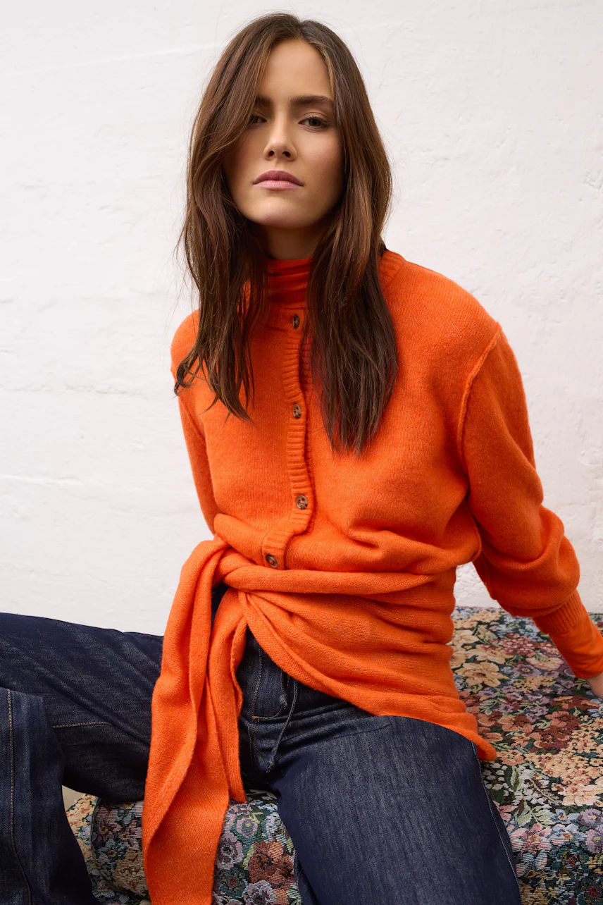 Woman in vibrant orange long-sleeve shirt, paired with dark jeans, sitting confidently on a patterned surface.