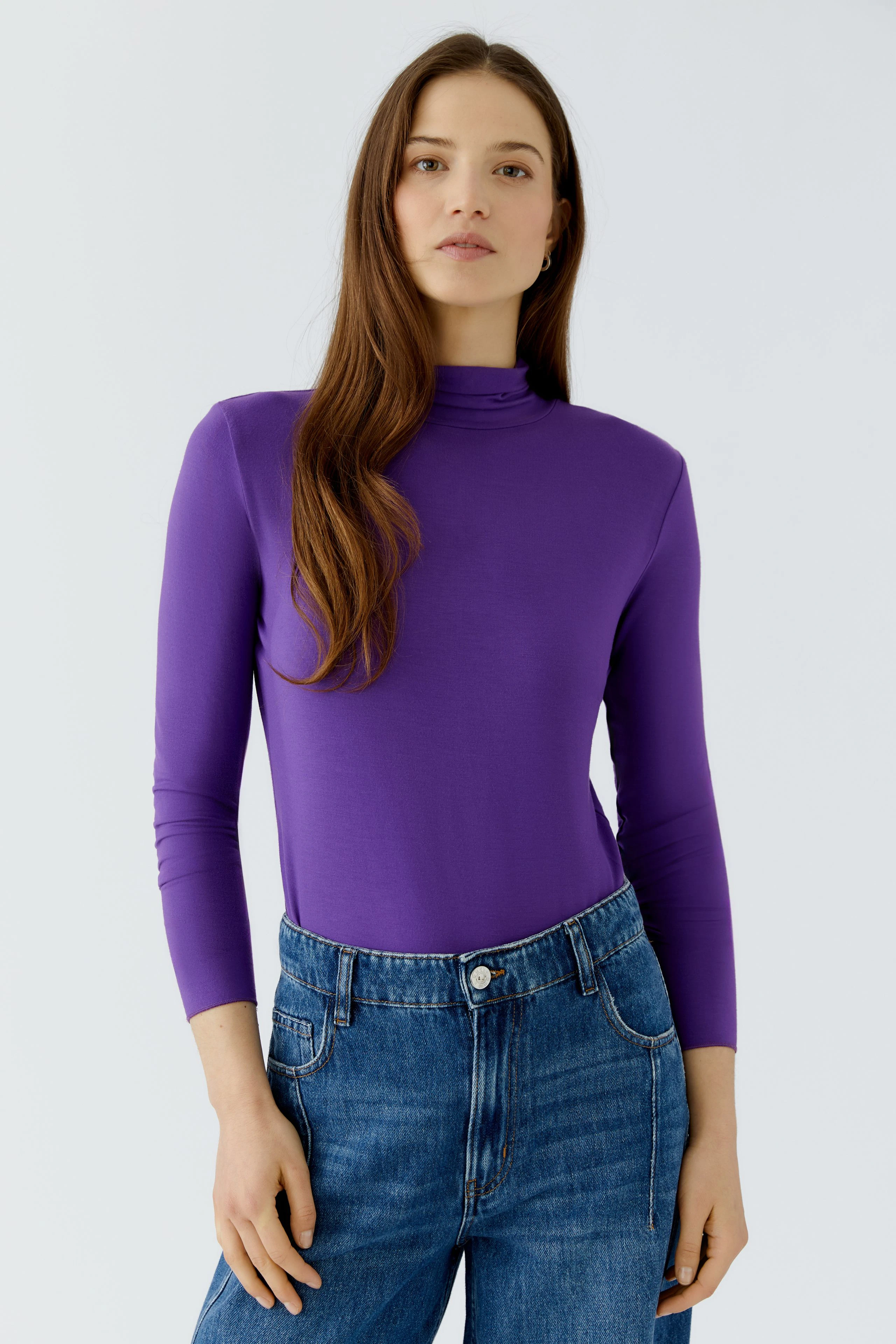 Casual long-sleeve shirt: Woman in purple top and blue jeans, standing confidently.