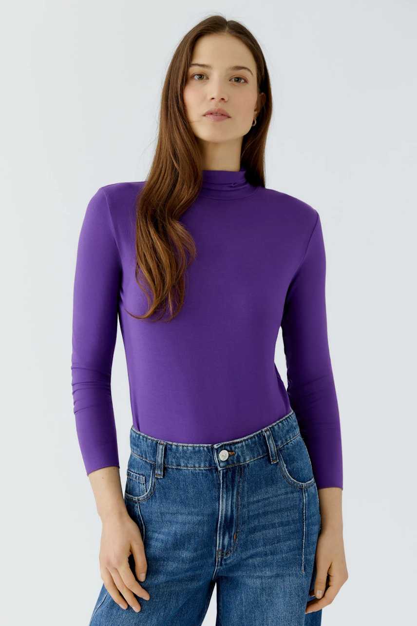 Casual long-sleeve shirt: Woman in purple top and blue jeans, standing confidently.