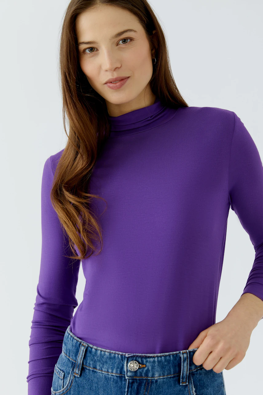 Woman in a purple long-sleeve shirt, paired with blue jeans, looking confidently at the camera.
