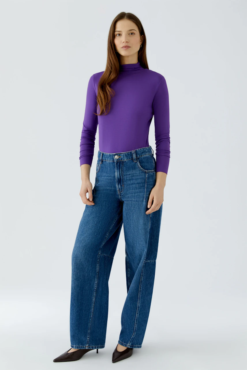 Woman in purple long-sleeve shirt and blue jeans, standing confidently.