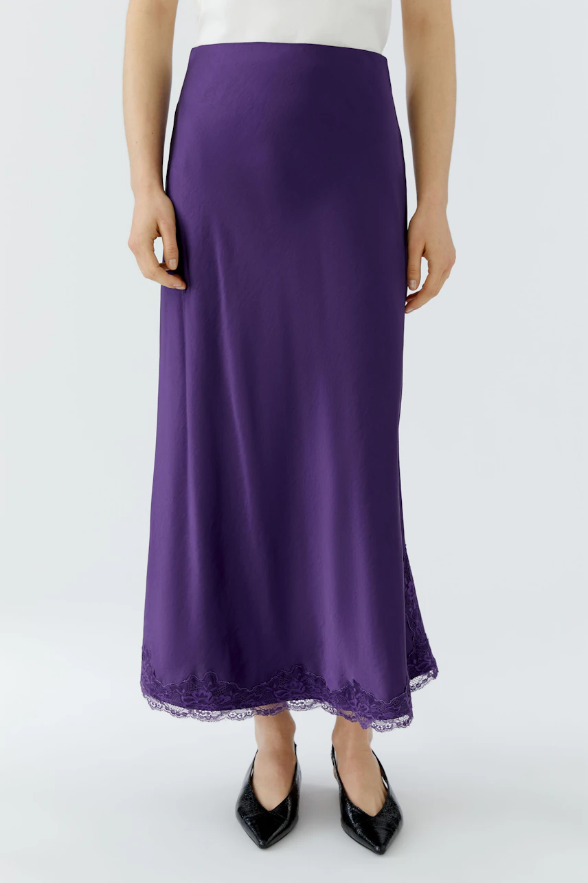 Woman in a purple maxi skirt with lace hem, paired with a white top and black shoes.