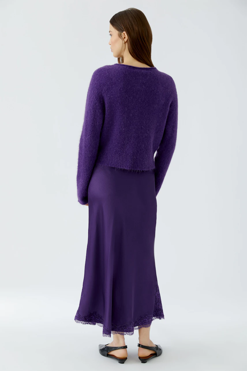Woman in purple maxi skirt and sweater, standing sideways. Skirt flows with lace hem, paired with black sandals.