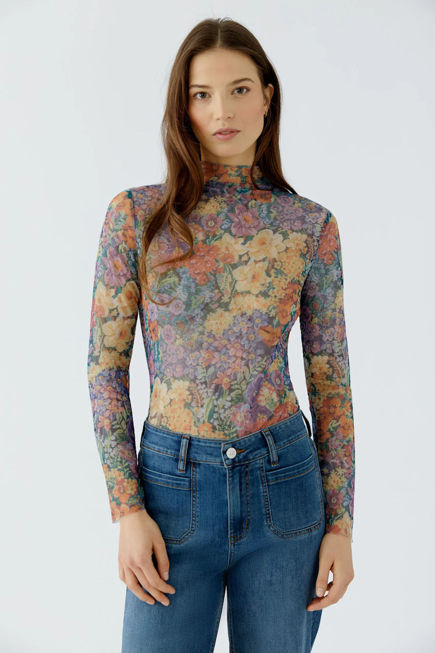 Frau im floralen Langarmshirt und Jeans, schaut direkt. Buntes Muster, schmaler Schnitt, lässiger Look.