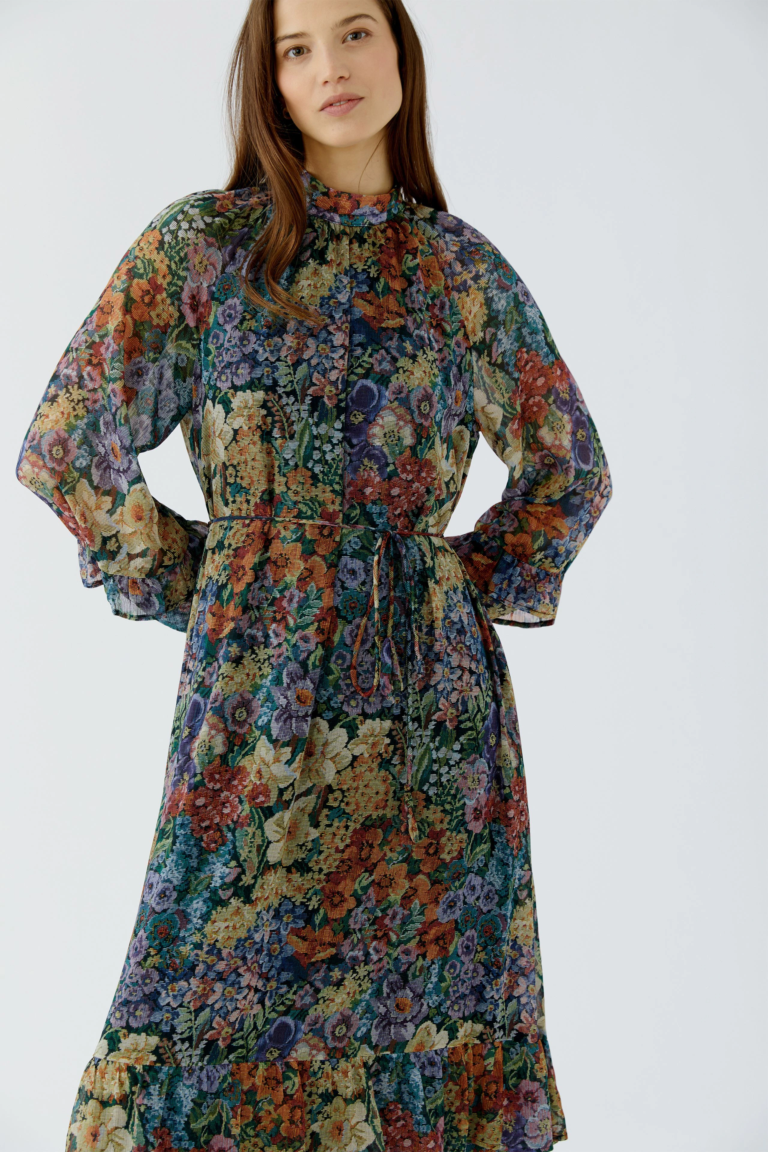 Frau im floralen Maxikleid mit langen Ärmeln und Stehkragen, blickt selbstbewusst.