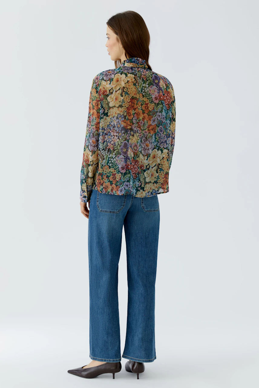 Frau in bunter Bluse mit Blumenmuster, kombiniert mit blauen Jeans und Absatzschuhen, von hinten gesehen.