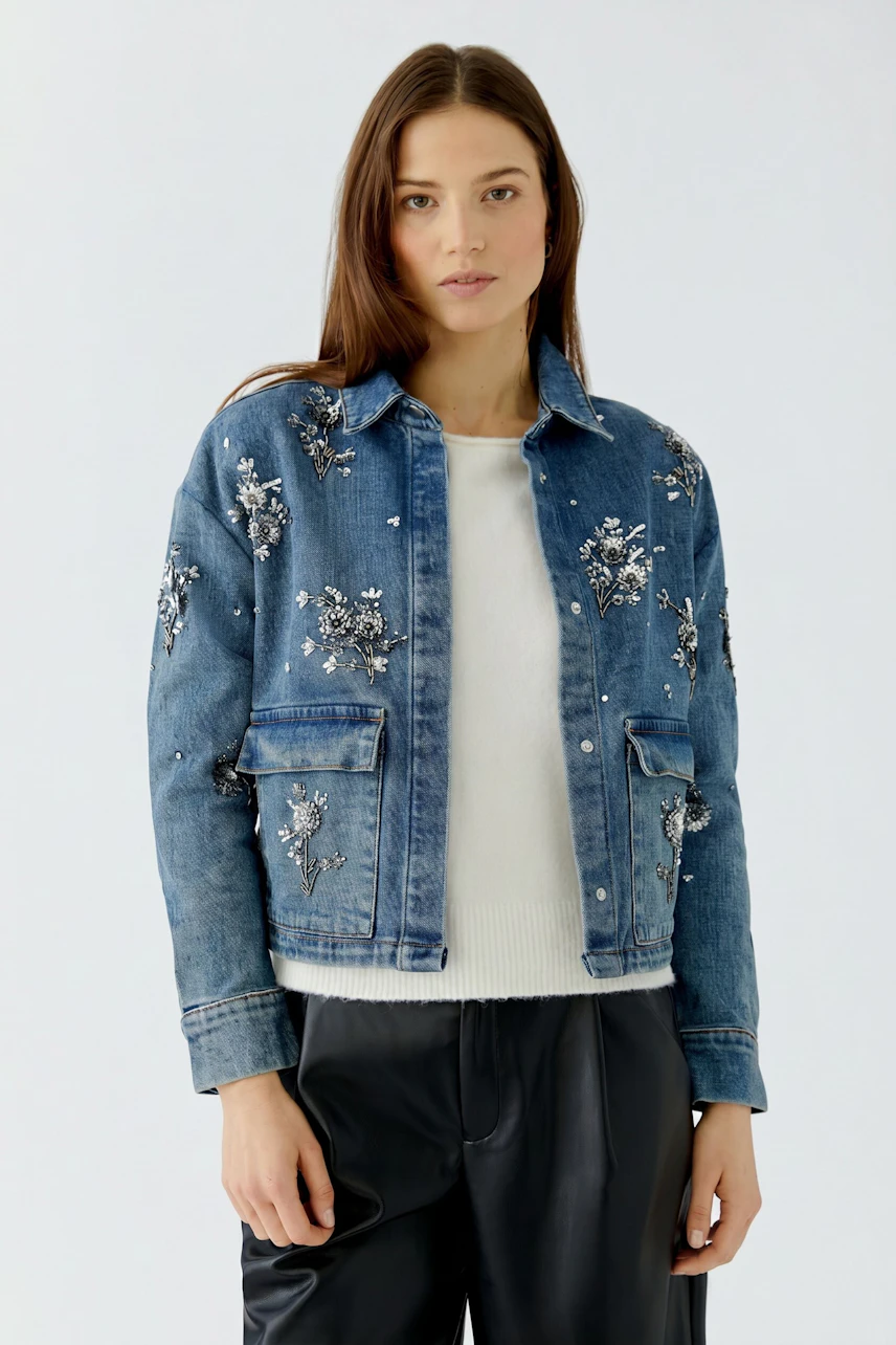 Woman in embroidered denim jacket over white pullover, paired with black pants, looking confidently.