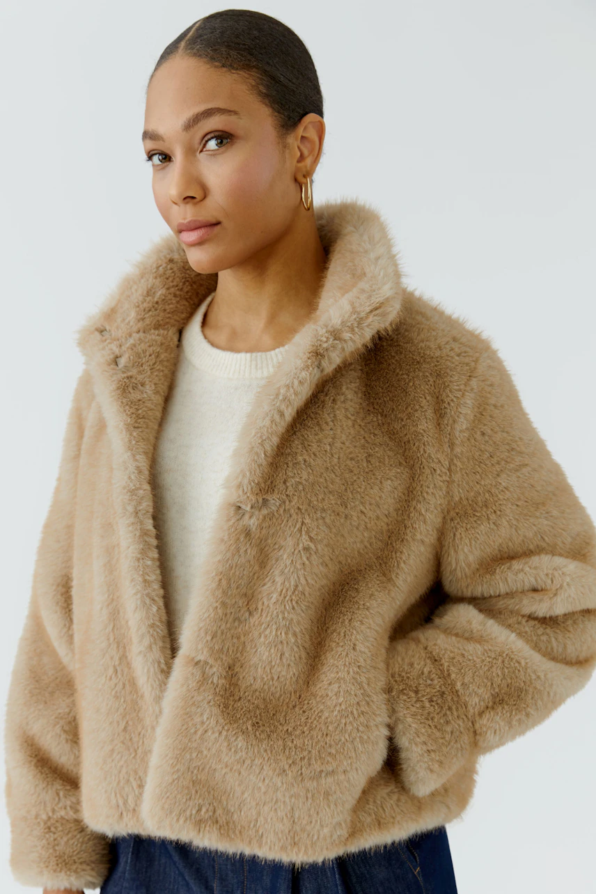 Woman in cozy outdoor jacket: Beige faux fur jacket over white pullover, hands in pockets, relaxed look.