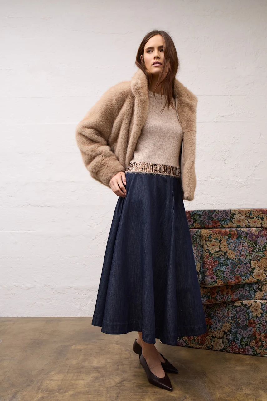 Woman in a cozy outdoor jacket, paired with a beige sweater and long denim skirt, standing confidently.