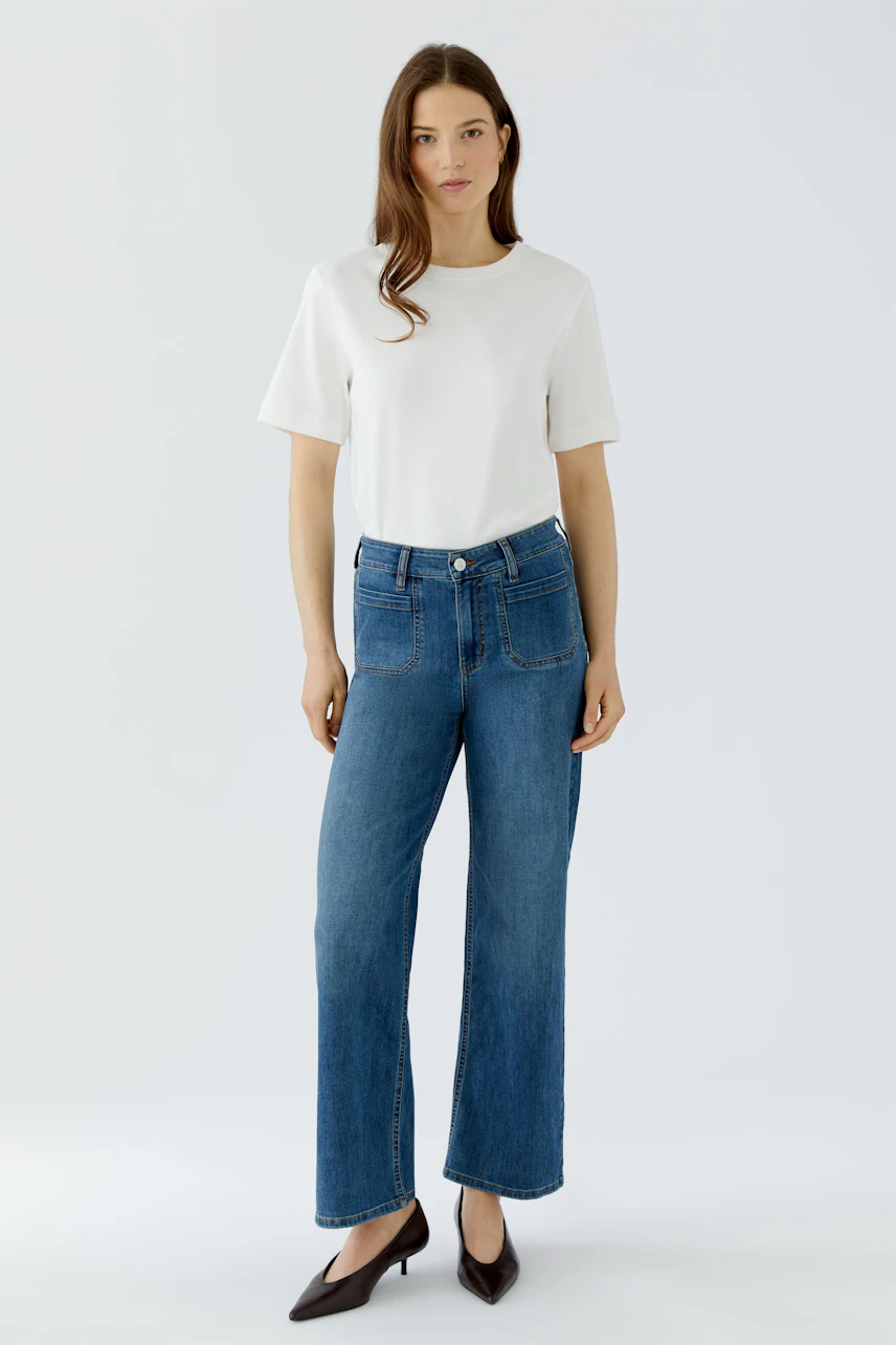 Frau in Jeans Straight und weißem T-Shirt, steht frontal, trägt schwarze Schuhe.