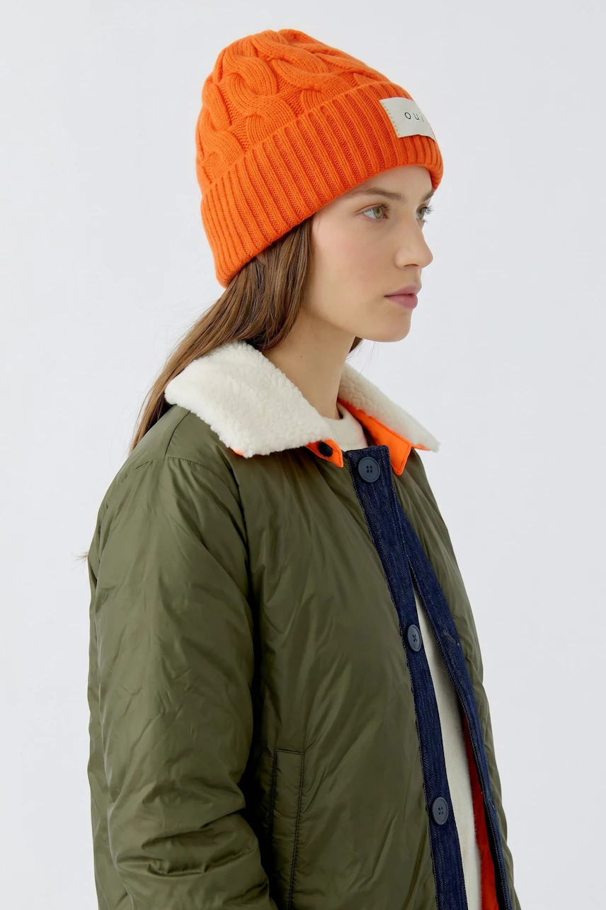 Frau mit orangefarbener Mütze und olivgrüner Jacke, seitlich blickend. Schlichtes, modernes Design.