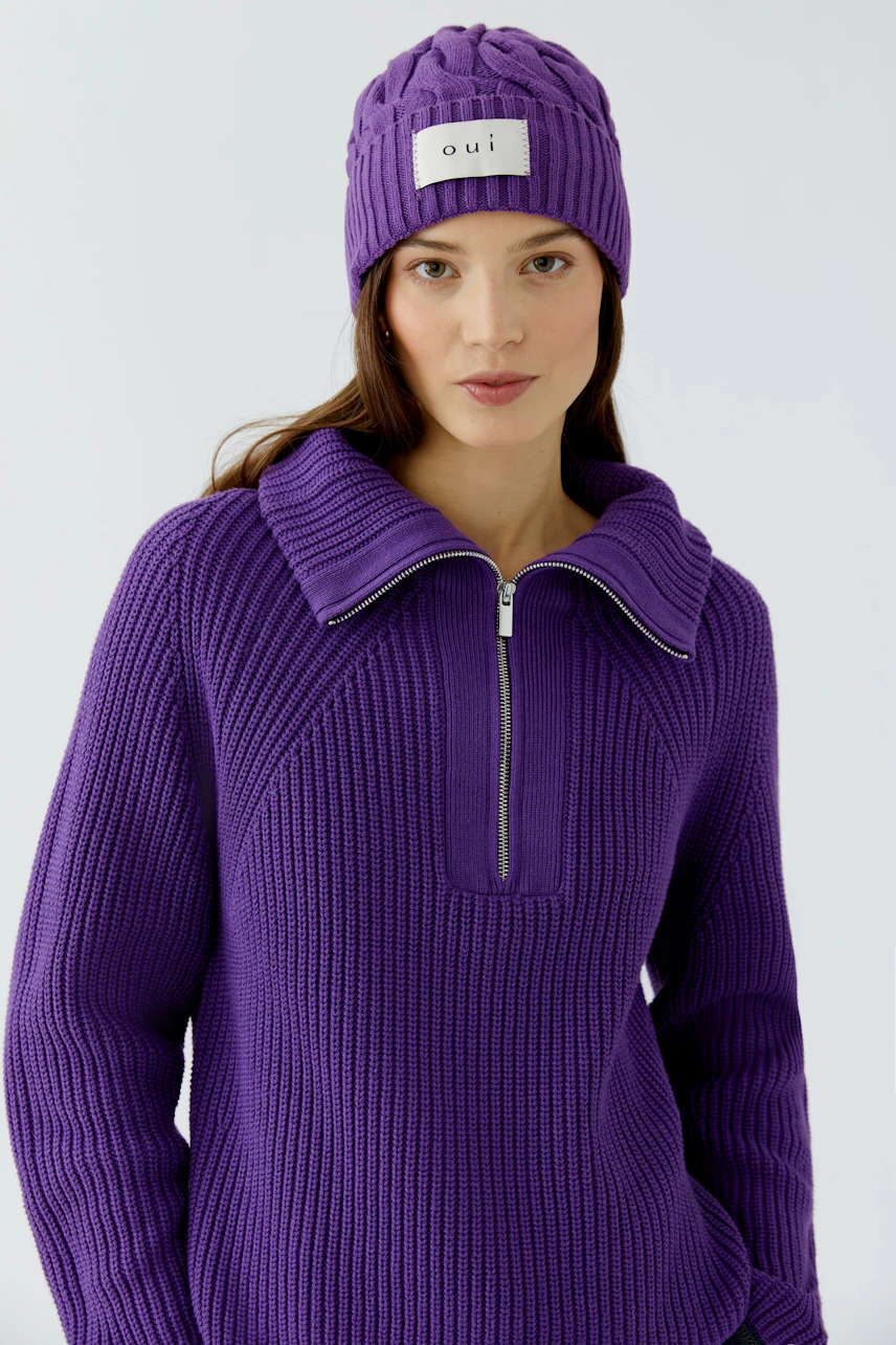 Frau mit lila Mütze und passendem Strickpullover, beide mit grobem Muster, schaut direkt in die Kamera.
