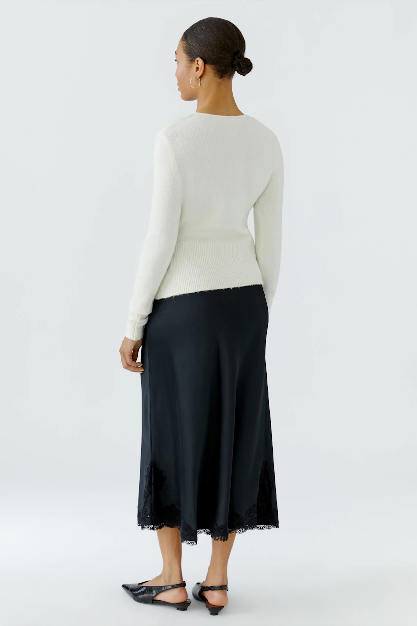 Woman in white cardigan and black skirt, standing confidently. Simple, stylish knitwear for everyday wear.