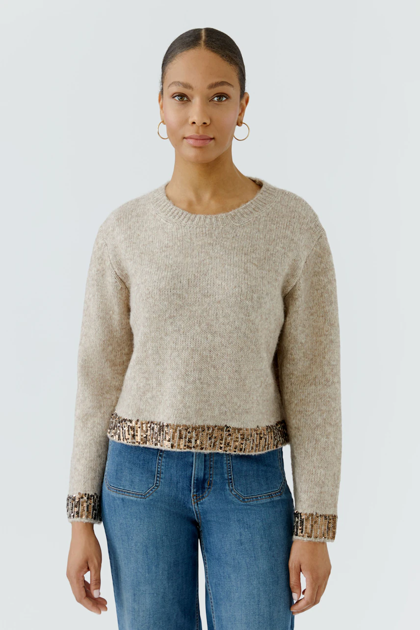 Casual women's pullover: Woman in beige pullover with sequin trim, paired with blue jeans, standing confidently.