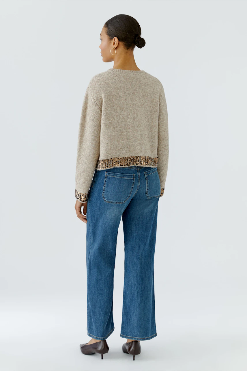 Casual women's pullover: Woman in beige pullover with patterned hem, paired with blue jeans, facing away.