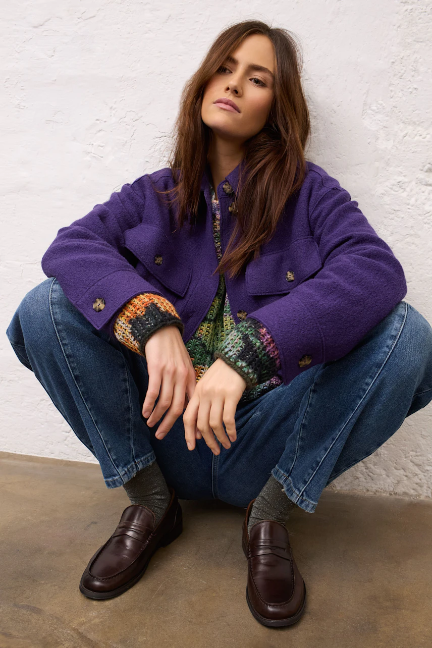 Casual overshirt: Woman in a purple overshirt, colorful pullover, and jeans, sitting confidently.