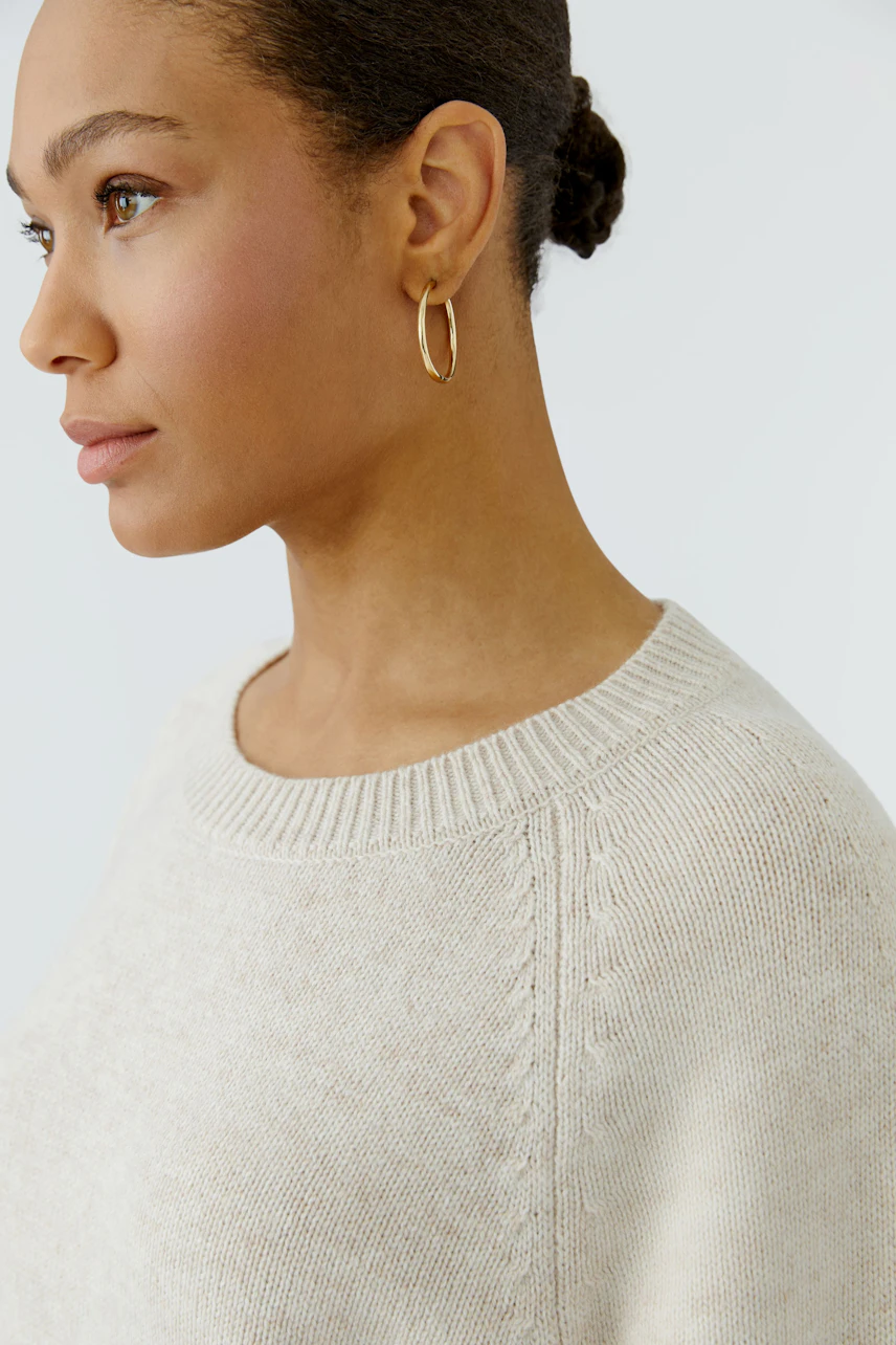 Frau im cremefarbenen Pullover mit Rundhalsausschnitt, schlichtes Design, goldene Ohrringe, seitliches Profil.